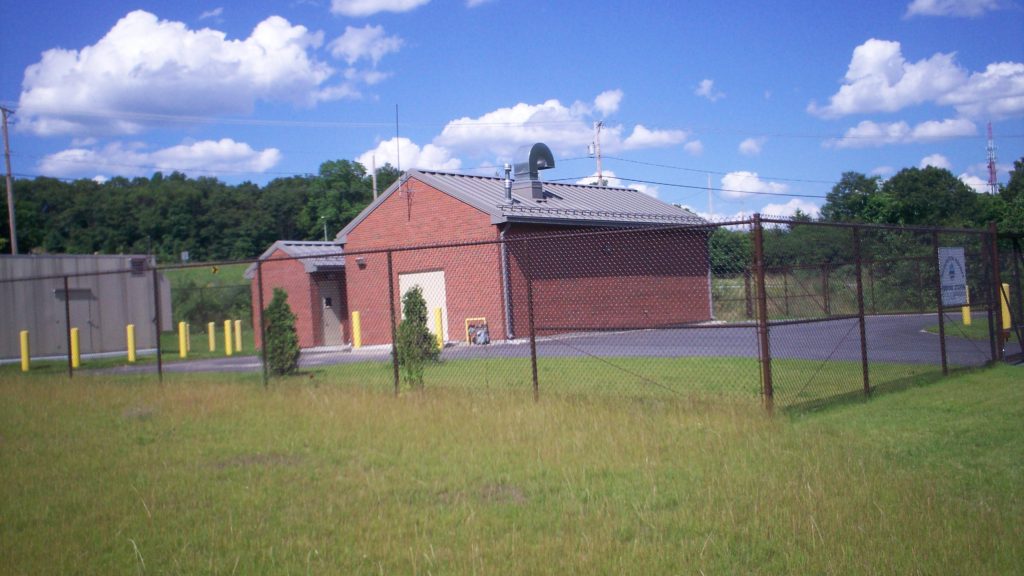 Exterior of a brick pump station building on a sunny day, enclosed with a chain link fence.