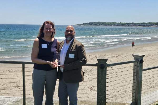 Engineer Nicole Iannuzzi and Joe Simmons stand side by side holding the ACEC-RI Engineering Excellence award. The background is a coastal beach on a sunny day.